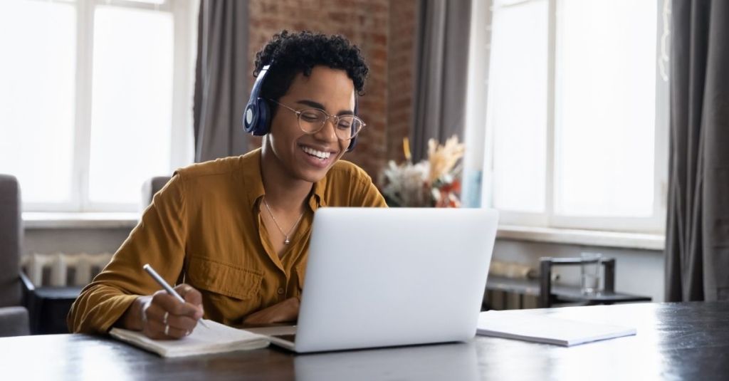A woman in a virtual meeting with her headset on while simultaneously writing on a notepad and laughing.